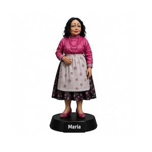 Puede incluir: Una figura de una persona con cabello oscuro ondulado, que viste una blusa magenta de manga larga, un delantal blanco con un pequeño patrón floral y una falda negra con estampado floral. La figura lleva zapatos rosas sobre una base negra Maria.