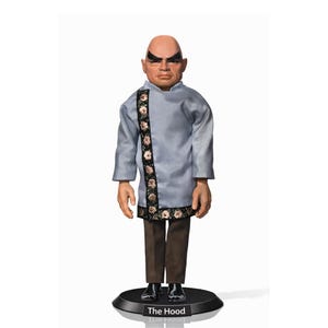 Puede incluir: Una figura de The Hood, un hombre calvo con una expresión severa, que lleva una túnica azul claro con adornos florales y pantalones marrones. La figura se encuentra sobre una base negra con el texto "The Hood".