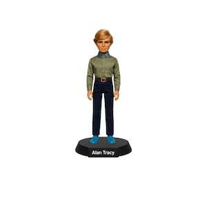 Puede incluir: Una figura coleccionable de Alan Tracy con cabello rubio, vistiendo una camisa de botones verde oliva de manga larga, un cinturón marrón con hebilla plateada, pantalones azul oscuro y zapatos azul brillante, sobre una base negra Alan Tracy.