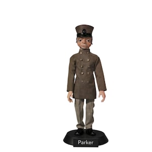 Puede incluir: Muñeca detallada de un hombre con uniforme y sombrero marrón, de pie sobre una base negra. El uniforme de la muñeca tiene varios botones y la base está etiquetada como "Parker". Los pantalones de la muñeca son a rayas.