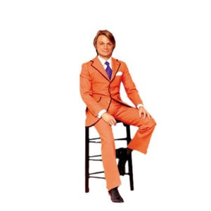 May include: A vintage orange suit with black trim, a white pocket square, and a purple tie. The suit is worn with black shoes. The person is seated on a black stool. The suit jacket has a two-button closure.