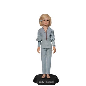 Pode incluir: Uma boneca da Lady Penelope em um terno azul claro com jaqueta e calças combinando. A boneca tem cabelo loiro e está em uma base preta com o texto "Lady Penelope". A jaqueta tem uma borda decorativa de contas.