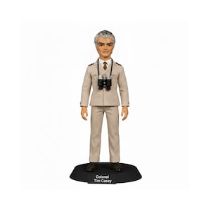May include: A standing figurine of Colonel Tim Casey wearing a beige military-style uniform with a white shirt, brown tie, and black binoculars around the neck. The figure is mounted on a black circular base with the name printed in white text.