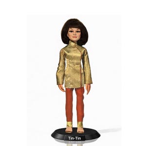 Puede incluir: Muñeca de Tin-Tin, vestida con una chaqueta dorada y pantalones color óxido. La muñeca tiene el pelo corto y castaño oscuro y está de pie sobre una base negra. La base tiene el nombre "Tin-Tin".
