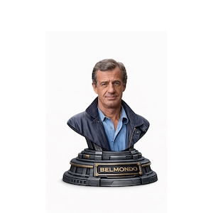 Peut inclure: Buste de Jean-Paul Belmondo, représentant la tête et les épaules de l'acteur. Il porte une chemise bleue et une veste en cuir bleu foncé. Le buste est monté sur un socle noir et or avec le nom "BELMONDO" affiché.