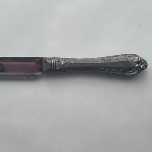 May include: A silver-colored bread knife with a decorative handle. The blade is dark and has a slightly curved edge. The handle features an ornate design. The knife is designed for cutting bread.