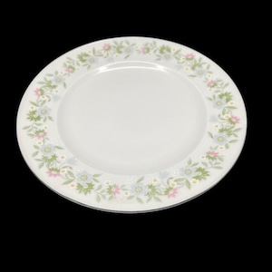 May include: White ceramic plate with a floral border featuring light blue, pink, and green flowers. The plate has a simple, elegant design, suitable for serving food. The plate is set against a black background.
