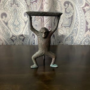 May include: A bronze-colored monkey figurine holding a rectangular tray above its head. The monkey is in a standing position with its arms raised. The figurine has a textured surface and is on a dark brown surface.
