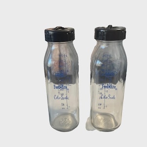 May include: Two clear glass baby bottles with black caps. The bottles have blue text that reads "DAVOL" and "Feed Rite Color Scale". The bottles have measurement markings.