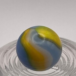 May include: A colorful glass marble with a swirling pattern of blue, yellow, and white. The marble is round and sits in a clear glass dish. The colors blend together, creating a unique and eye-catching design.