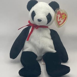May include: A plush panda bear with black and white fur, a red ribbon tied around its neck, and a heart-shaped tag. The panda has black eyes and ears. The stuffed animal is sitting upright.