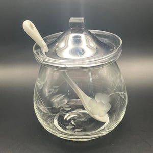 May include: A clear glass sugar bowl with a silver lid and a white spoon. The bowl has etched floral designs. The spoon rests inside the bowl, and the lid is slightly ajar.