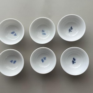 May include: Six small, white ceramic bowls with a simple, rounded design. Each bowl features a blue, stylized character in the center. The bowls are arranged in two rows of three on a light gray surface.