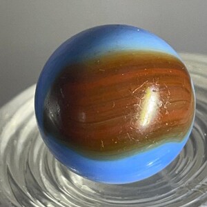 May include: A glass marble with a blue and brown swirl pattern. The marble has a glossy finish and is resting on a clear, ridged surface. The colors blend together, creating a unique and eye-catching design.