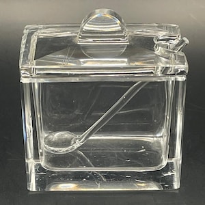 May include: A clear, square-shaped glass sugar bowl with a lid and a small spoon. The lid has a rounded handle. The spoon rests inside the bowl. The glass is transparent, allowing the contents to be visible.