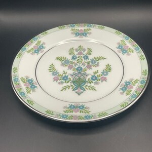 May include: White ceramic plate with a floral design in blue, pink, and green. The plate has a silver rim and a light green border. The floral pattern is centered on the plate and repeated around the edge.