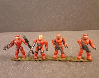 Peut inclure: Quatre figurines rouges de soldats, chacune avec une arme différente. Les figurines sont sur une base verte. Une figurine a un casque doré. Les figurines sont alignées, sur fond gris.