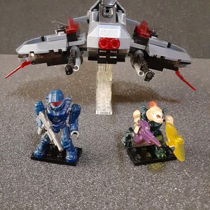 May include: A gray and red spaceship with two blue and silver figures standing in front of it. The figures are holding weapons.