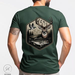 Puede incluir: Camiseta verde oscuro con un diseño temático de la naturaleza. El diseño incluye un ciervo, un pez, un pato y árboles, todo dentro de un marco hexagonal. La obra de arte utiliza tonos marrones, negros y blancos, creando una estética rústica.