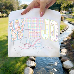 Può includere: Felpa bianca con la scritta "WWJD" in lettere colorate e decorate. La "W" è floreale, la seconda "W" è a quadri e la "JD" ha croci e fiocchi. Un disegno di fiocco pastello è sotto il testo.