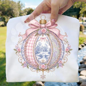 May include: A white t-shirt featuring a graphic of a decorative Christmas ornament. The ornament has a pink gingham section, a blue and white toile landscape scene, and a paisley pattern, all accented with pink bows, roses, and gold filigree.