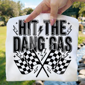 May include: A white t-shirt featuring a black graphic print with the text "HIT THE DANG GAS" in a bold, distressed font. The design includes two crossed checkered racing flags and several lightning bolt icons surrounding the text.