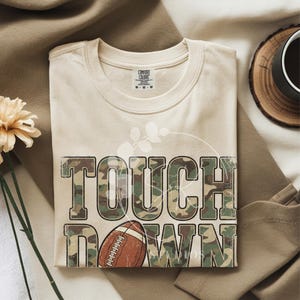 May include: A beige short-sleeved t-shirt featuring the text TOUCH DOWN in a green, brown, and tan camouflage pattern. The letter O in DOWN is replaced by a brown and white football graphic. A faint white leaf outline is visible behind the text.