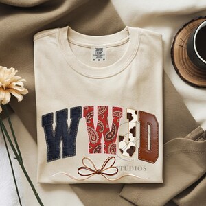 Może przedstawiać: Kremowa bluza z literami "WWD" w patchworkowym wzorze. "W" jest dżinsowe, drugie "W" jest czerwone paisley, "D" jest ze skóry brązowej, a "D" ma nadruk w krowy. Brązowa kokarda i słowo "STUDIOS" znajdują się poniżej.