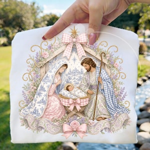 May include: A white fabric item featuring a printed nativity scene illustration. The design shows a figure in a blue and white patterned cloak and a figure in a blue plaid cloak kneeling beside a baby in a manger, framed by floral garlands and bows.