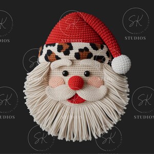 Può includere: Un volto di Babbo Natale all'uncinetto con cappello rosso, fascia leopardata e barba bianca. Il volto presenta naso rosso, guance rosa e bocca rossa. Lo sfondo è nero.