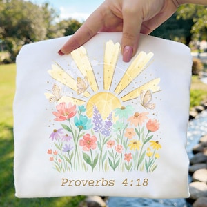 Op de afbeelding: Een wit t-shirt met een afbeelding van een gele zon met stralende zonnestralen, twee vlinders en een kleurrijk veld met aquarel-stijl bloemen in tinten roze, blauw, paars en geel, met de tekst Proverbs 4:18 eronder gedrukt.
