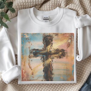 May include: White crewneck sweatshirt featuring a painted cross design. The cross is brown with gold accents, set against a backdrop of blue, pink, and gold brushstrokes. The sweatshirt has a classic, comfortable fit.