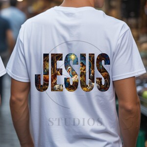 May include: A white short-sleeved t-shirt featuring the word JESUS in large block letters on the back. The letters contain a detailed nativity scene illustration depicting Mary, Joseph, the infant Jesus, a star, and various animals in a stable.