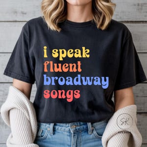 May include: Black t-shirt with the text "i speak fluent broadway songs" in yellow, orange, blue, and red. The shirt is a casual style, suitable for everyday wear. The design is a playful statement for musical theater enthusiasts.