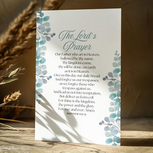 May include: A white card with a blue and green watercolour floral design. The card features the Lord's Prayer with the text "The Lord's Prayer" in a large, decorative font. The text includes the words "Our Father who art in Heaven, hallowed be thy name." and "Matthew 6:9-13".