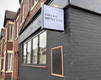 Pode incluir: Um edifício de tijolo preto com uma fachada de loja que apresenta uma placa branca que diz "Cafe Bar Bottle Shop". A fachada tem grandes janelas e um toldo preto com a palavra "TRATTORIA" escrita nele.