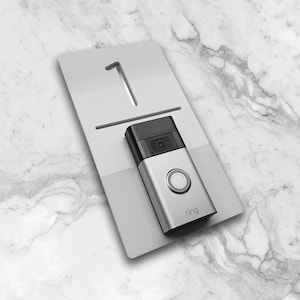 May include: A silver Ring Video Doorbell with a black camera lens and a white button is mounted on a white plastic plate with the number "1" printed on it.