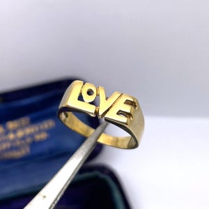 May include: A gold-toned ring featuring the word LOVE as the centrepiece, with the letters formed by the metal band itself. The ring has a polished finish and is held by a metal tool, showcasing the cutout design of the letters L, O, V, and E.