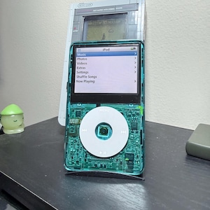 Puede incluir: Un iPod turquesa con una rueda de control blanca y una pantalla que muestra las opciones del menú: Música, Fotos, Vídeos, Extras, Ajustes, Reproducción aleatoria y Reproduciendo ahora. Detrás del iPod hay una consola de juegos portátil retro.