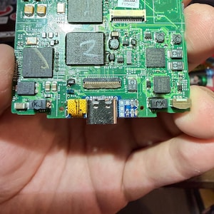 Puede incluir: Una placa de circuito verde con varios componentes electr&oacute;nicos, incluyendo chips, resistencias y conectores. La placa tiene texto que dice "APPLE" y "2005". La placa se sostiene en una mano.