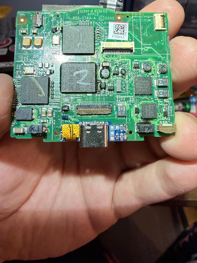 Puede incluir: Una placa de circuito verde con varios componentes electr&oacute;nicos, incluyendo chips, resistencias y conectores. La placa tiene texto que dice "APPLE" y "2005". La placa se sostiene en una mano.