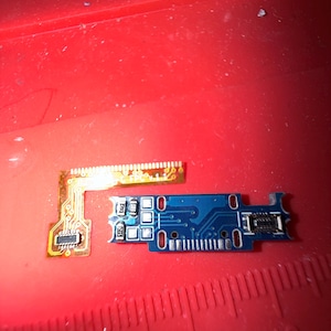 Puede incluir: Primer plano de dos placas de circuito electr&oacute;nico. Una es una peque&ntilde;a placa azul rectangular con varios agujeros y componentes. La otra es una placa de circuito flexible delgada y dorada. Ambas est&aacute;n sobre una superficie roja.