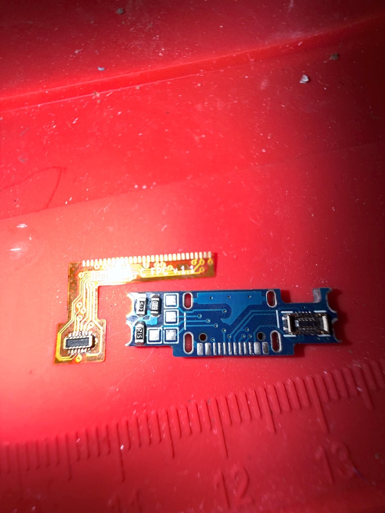 Puede incluir: Primer plano de dos placas de circuito electr&oacute;nico. Una es una peque&ntilde;a placa azul rectangular con varios agujeros y componentes. La otra es una placa de circuito flexible delgada y dorada. Ambas est&aacute;n sobre una superficie roja.