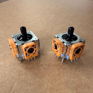 May include: Two orange and silver joystick modules with black joysticks. Each module has a square base with orange side panels and a black joystick extending upwards. The modules are designed for electronic devices.
