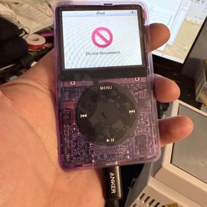 Puede incluir: Un iPod morado transparente con una rueda de clic negra. La pantalla muestra un s&iacute;mbolo de prohibici&oacute;n rojo y el texto "Do not disconnect". Un cable de carga Anker est&aacute; conectado.