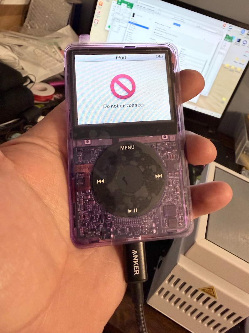 Puede incluir: Un iPod morado transparente con una rueda de clic negra. La pantalla muestra un s&iacute;mbolo de prohibici&oacute;n rojo y el texto "Do not disconnect". Un cable de carga Anker est&aacute; conectado.