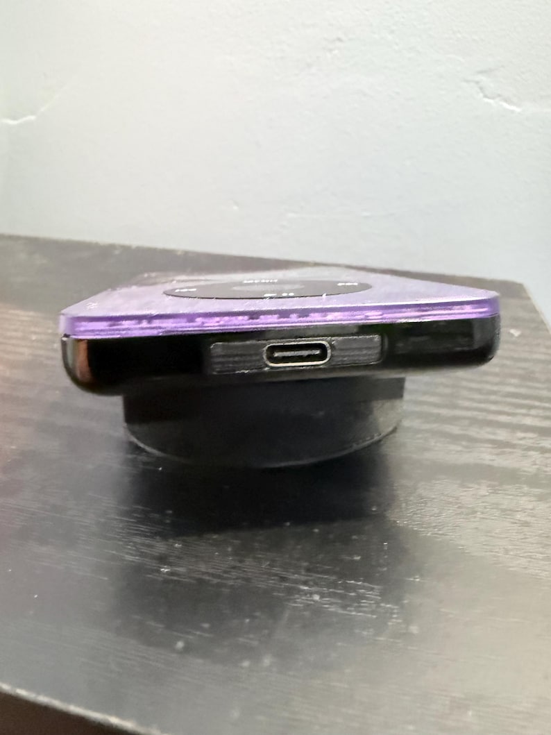 Puede incluir: Un dispositivo electr&oacute;nico negro y morado, posiblemente un tel&eacute;fono o reproductor multimedia, descansa sobre una base de carga inal&aacute;mbrica negra. El dispositivo tiene una carcasa morada y negra. La base de carga es redonda y negra. La imagen est&aacute; tomada desde un &aacute;ngulo lateral.