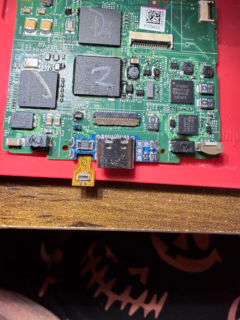 Puede incluir: Primer plano de una placa de circuito con varios componentes electr&oacute;nicos. La placa es principalmente verde con detalles en rojo y dorado. Se ve un puerto USB-C, junto con varios chips y conectores. La imagen est&aacute; bien iluminada.