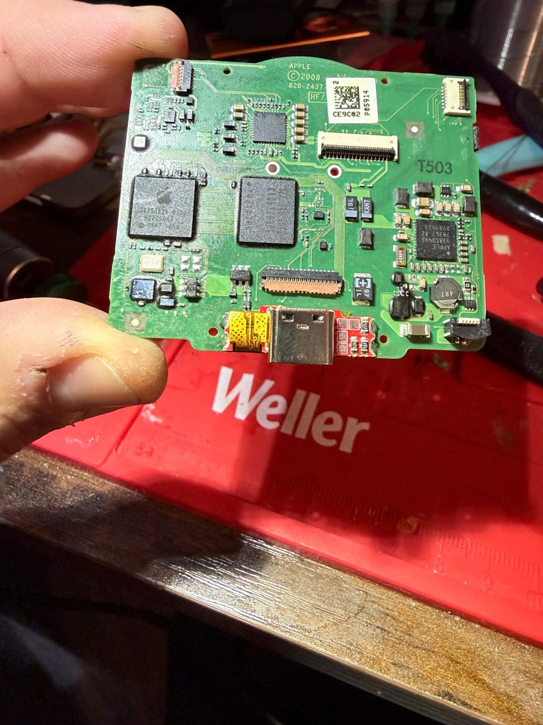 May include: A green circuit board with various electronic components, including chips, connectors, and a USB-C port. The board has the Apple logo and the text "APPLE &copy;2008 820-2437". The board is held by a person's fingers.