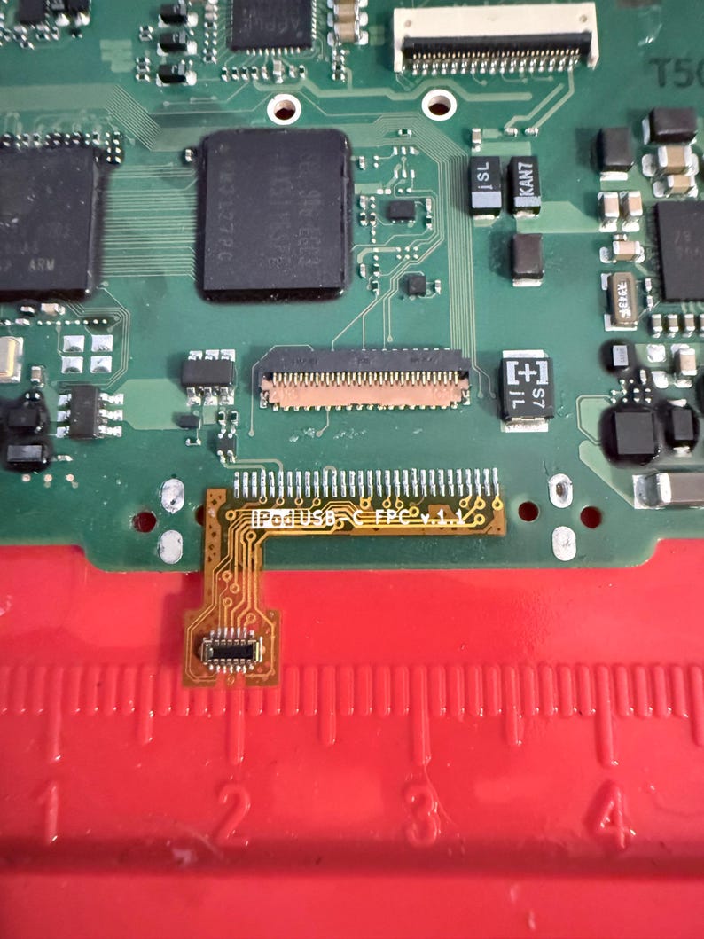 May include: A close-up of a green circuit board with various electronic components. A small, flexible circuit board labeled "iPodUSB FPC v.1.1" is connected to the main board. A red ruler is visible at the bottom, with markings in inches.